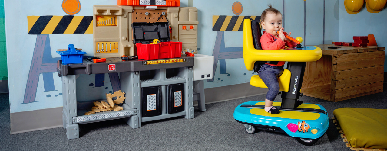 A young toddler sitting in a Permobile Explorer Mini in a playroom where the child can approach a toy workbench